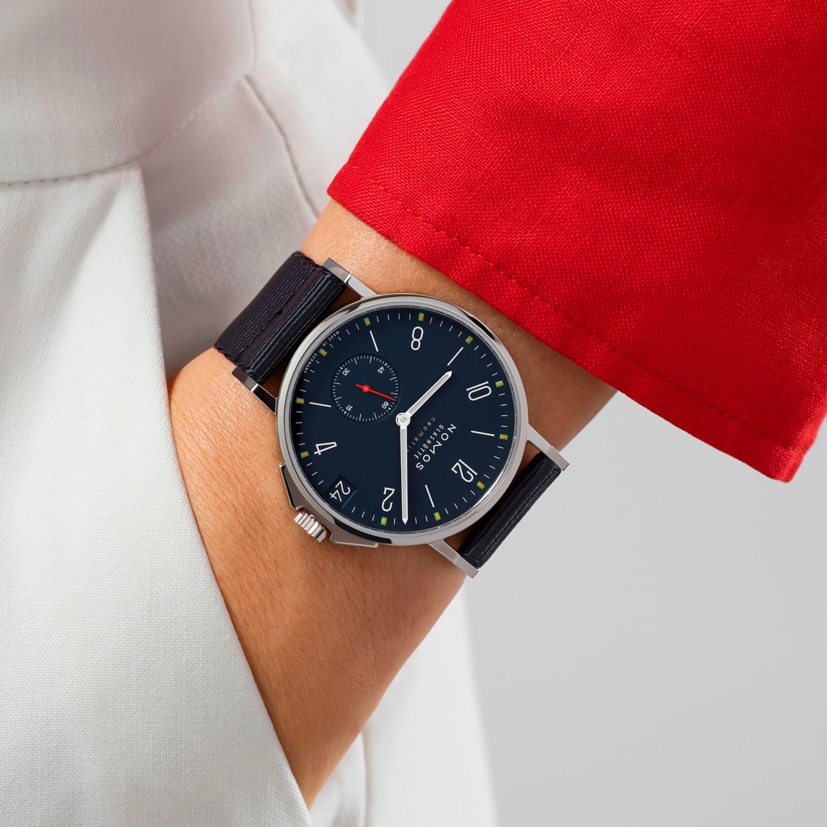 2025 © NOMOS GLASHÜTTE Eine Person trägt eine silberne NOMOS Glashütte Ahoi-Armbanduhr mit schwarzem Armband und marineblauem Zifferblatt mit weißen Ziffern, dazu ein weißes Outfit mit rotem Ärmel.