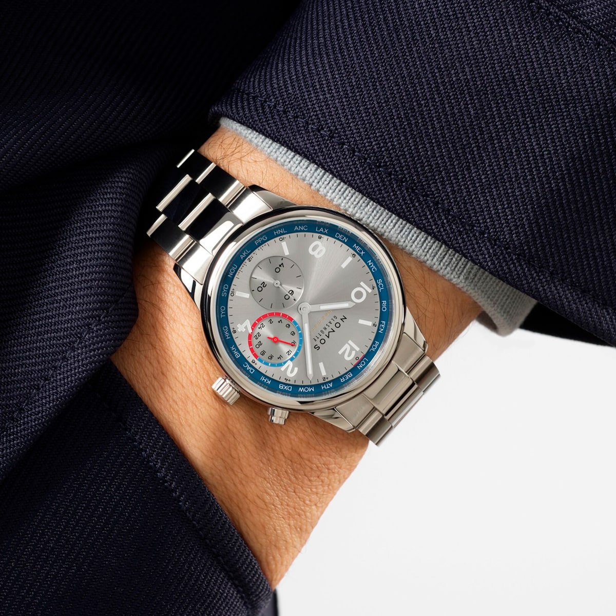 2025 © NOMOS GLASHÜTTE Eine Person in einem marineblauen Anzug zeigt eine NOMOS Glashütte Club-Armbanduhr mit blauer Lünette, grauem Zifferblatt, silbernen Indexen, Hilfszifferblättern und einem rot akzentuierten Bereich. Der Zeiger ist in der Anzugtasche verstaut.