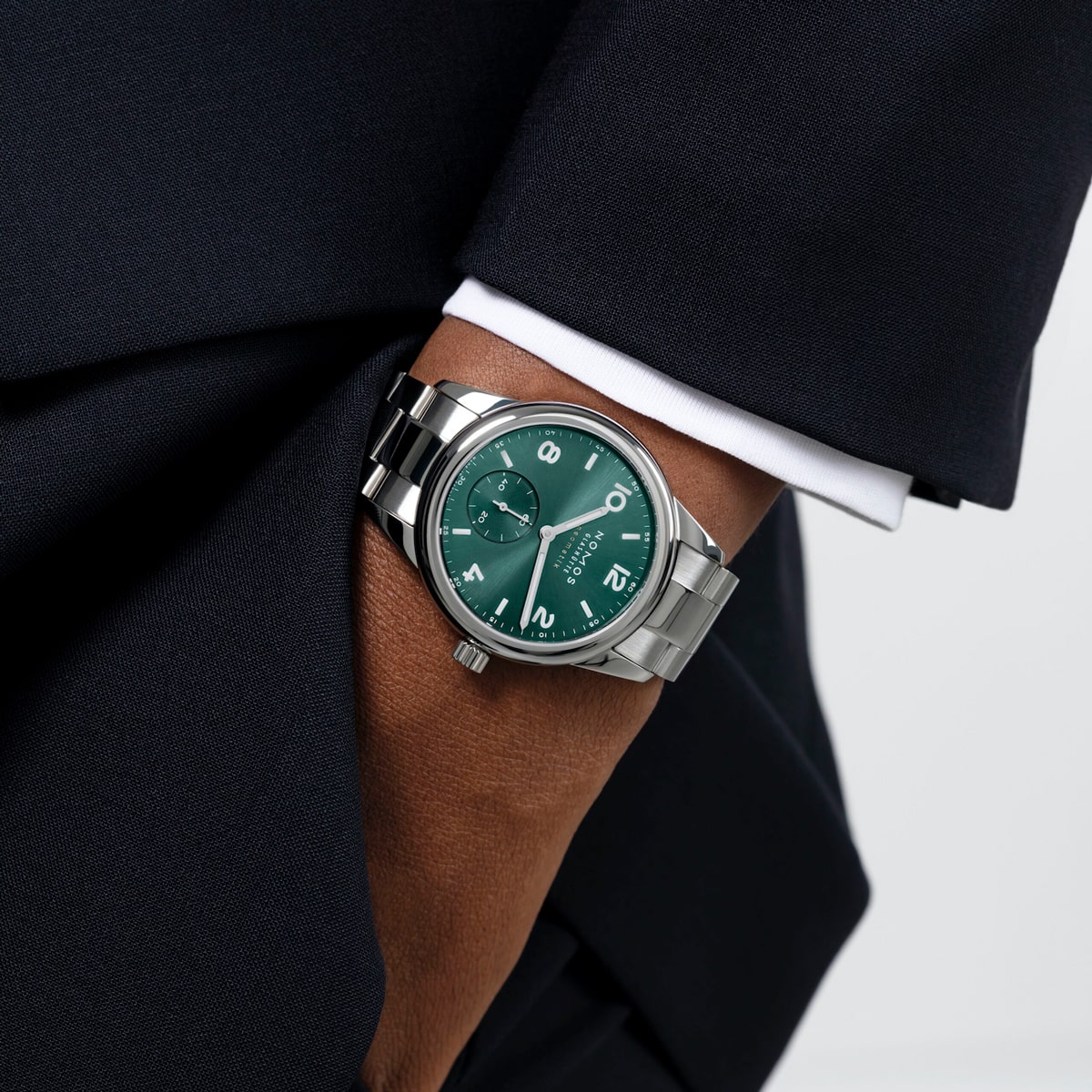 2025 © NOMOS GLASHÜTTE Eine Person im dunklen Anzug hat die Hand in der Tasche und zeigt eine silberne NOMOS Glashütte Club-Armbanduhr mit grünem Zifferblatt und weißen Ziffern.