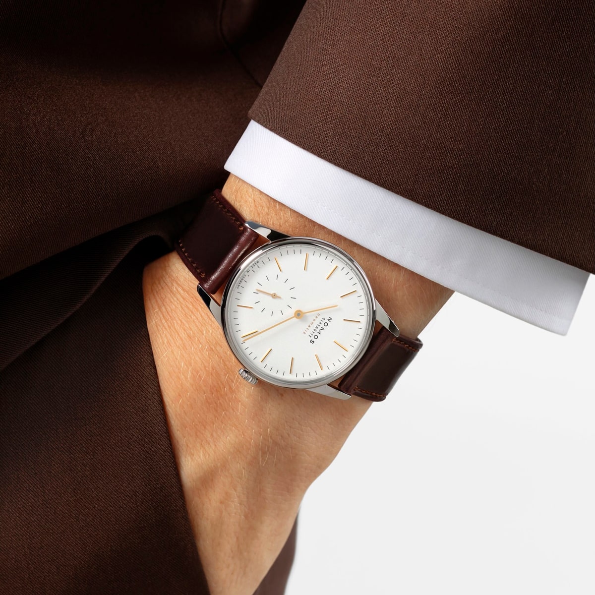 2025 © NOMOS GLASHÜTTE Eine Person in braunem Anzug und weißem Hemd trägt eine NOMOS Glashütte Orion mit weißem Zifferblatt und braunem Lederband am Handgelenk. Die silberne Uhr zeigt die Zeit von etwa 3:43 Uhr an.