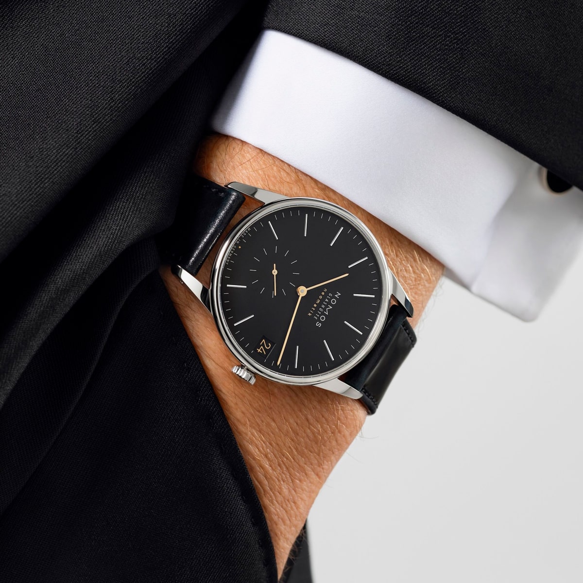 2025 © NOMOS GLASHÜTTE Eine Person in schwarzem Anzug und weißem Hemd zeigt eine NOMOS Glashütte Orion-Armbanduhr mit schwarzem Lederarmband und schwarzem Zifferblatt, mit minimalistischen Stundenmarkierungen und kleiner Sekunde.