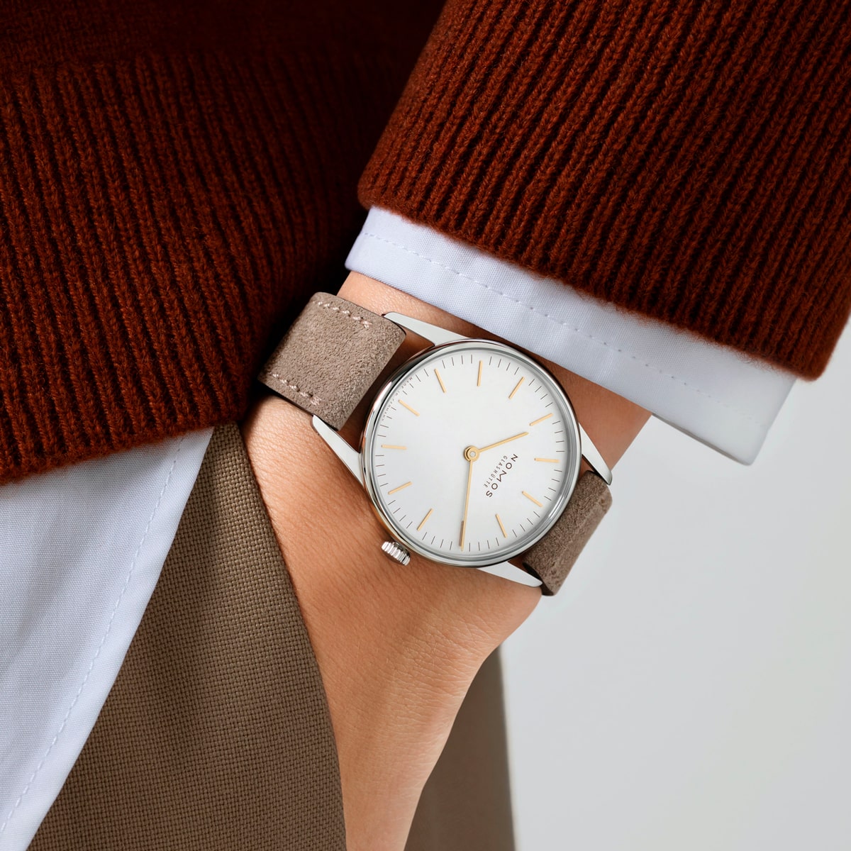2025 © NOMOS GLASHÜTTE Eine Person, die einen braunen Pullover und eine beigefarbene Hose trägt, hat eine minimalistische silberne Armbanduhr NOMOS Glashütte Orion mit weißem Zifferblatt und hellbraunem Lederband am linken Handgelenk.