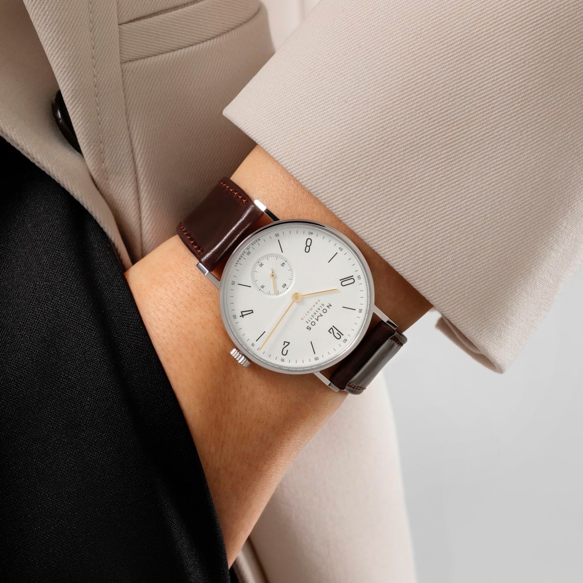 Ein Träger eines beigen Blazers zeigt eine NOMOS Glashütte Tangente-Armbanduhr mit weißem Zifferblatt, schwarzen Ziffern, kleiner Sekunde und braunem Lederband.
