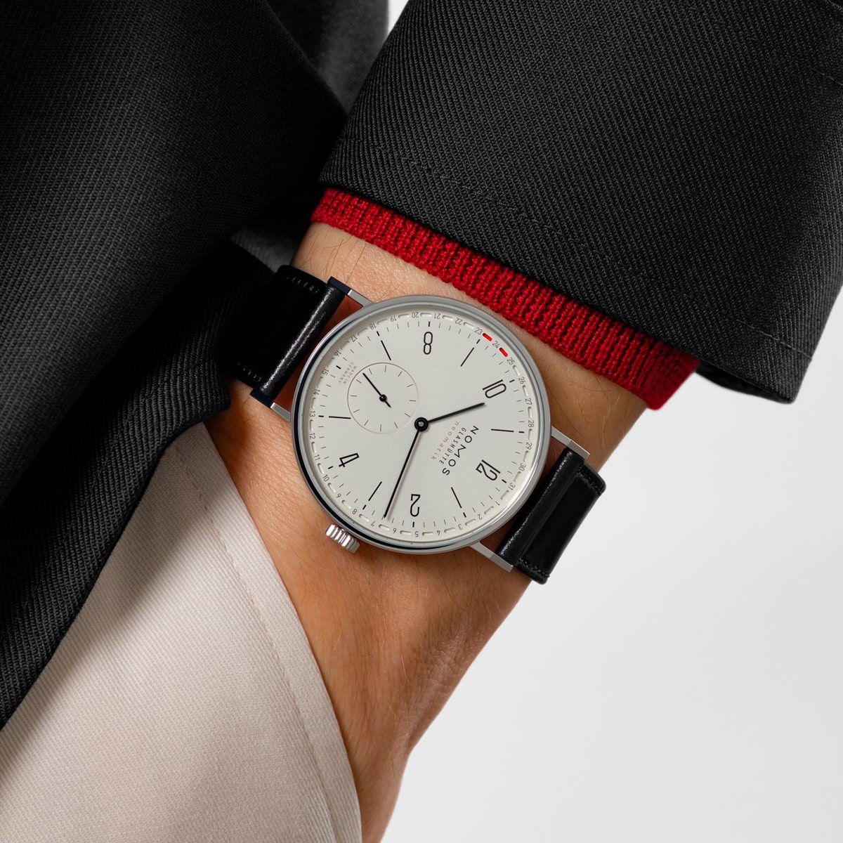Eine Person im schwarzen Anzug mit roter Manschette zeigt eine minimalistische NOMOS Glashütte Tangente-Armbanduhr mit weißem Zifferblatt, schwarzem Lederband und schlichten schwarzen Ziffern und Zeigern.