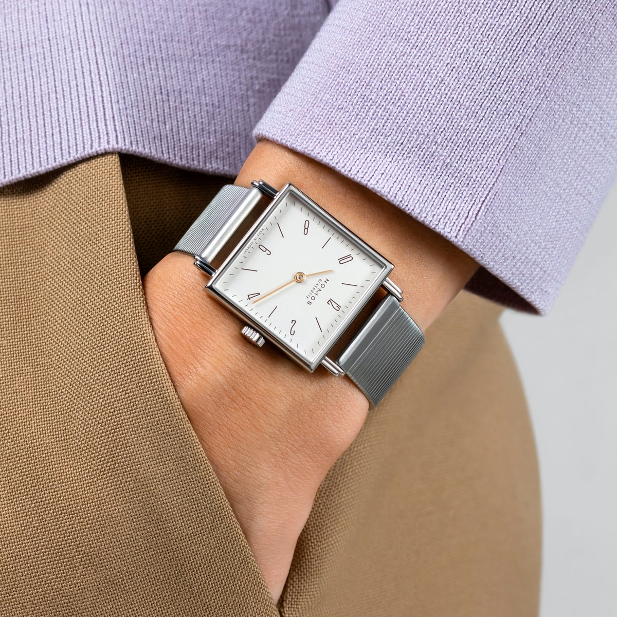 2025 © NOMOS GLASHÜTTE Eine Person, die eine silberne NOMOS Glashütte Tetra mit quadratischem Zifferblatt und Metallband trägt, bekleidet mit einem lavendelfarbenen Pullover und einer hellbraunen Hose, hat die Hand in der Tasche.