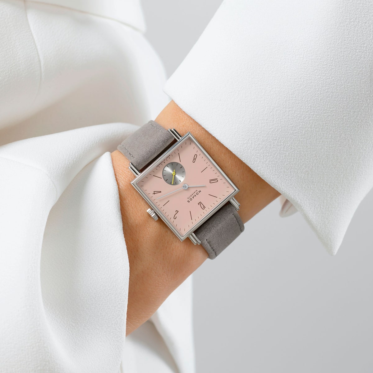 2025 © NOMOS GLASHÜTTE Eine Person, die ein weißes langärmeliges Kleidungsstück trägt, zeigt eine NOMOS Glashütte Tetra-Armbanduhr mit rosafarbenem Zifferblatt, minimalen Markierungen und einem grauen Wildlederarmband. Die Uhr verfügt über ein kleines Hilfszifferblatt und hat ein schlichtes, modernes Design.