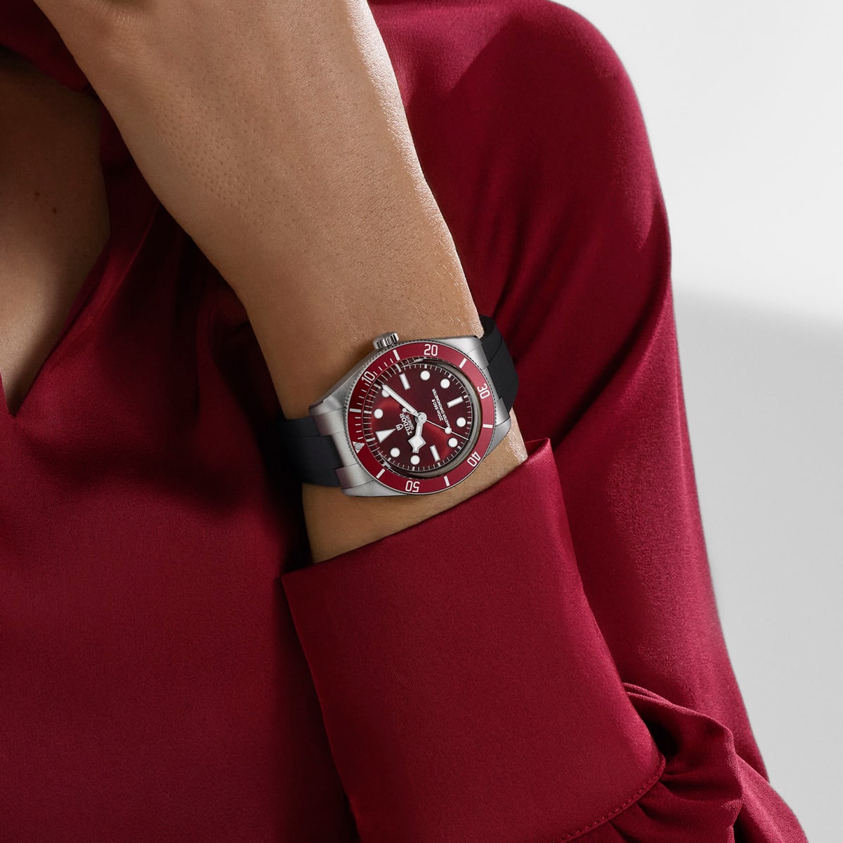 2025 © TUDOR Eine Person, die ein langärmeliges, dunkelrotes Hemd trägt, hat eine silberne Black Bay 58-Armbanduhr mit rotem Ziffernblatt und schwarzem Armband am Handgelenk, die Hand ruht in der Nähe ihres Kinns.