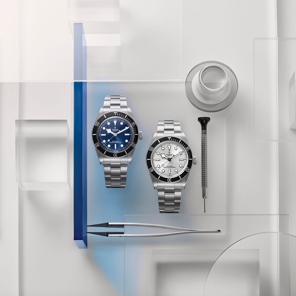 2025 © TUDOR Zwei Luxusarmbanduhren, darunter eine Black Bay 68 mit Metallband, ein Schraubenzieher, eine Pinzette und eine Lupe, sind auf einer hellen, geometrischen Fläche ausgestellt. Die linke Uhr hat ein blaues Zifferblatt, die rechte Uhr ein weißes.