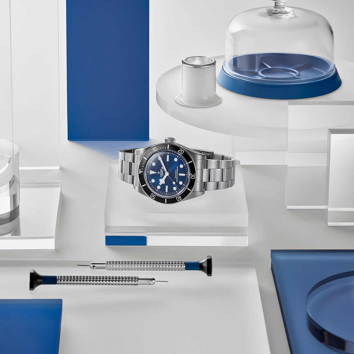 2025 © TUDOR Eine silberne Armbanduhr mit blauem Zifferblatt, die von der Black Bay 68 inspiriert ist, wird auf einer durchsichtigen Plattform präsentiert, umgeben von Uhrmacherwerkzeugen, einer Lupe und blauen und weißen geometrischen Objekten in einem minimalistischen Rahmen.