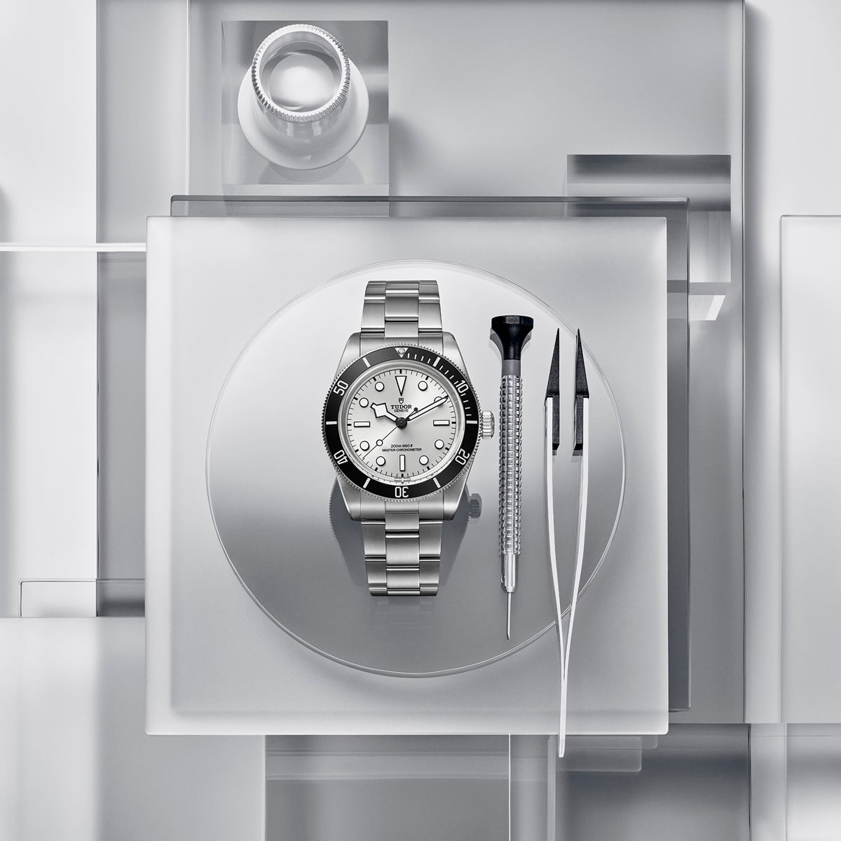 2025 © TUDOR Eine silberne Black Bay 68-Armbanduhr wird auf einem runden Tablett präsentiert, begleitet von einem Schraubenzieher und einer Pinzette. Der Hintergrund besteht aus übereinander geschichteten transparenten Quadraten, die einen modernen, minimalistischen Look vermitteln.