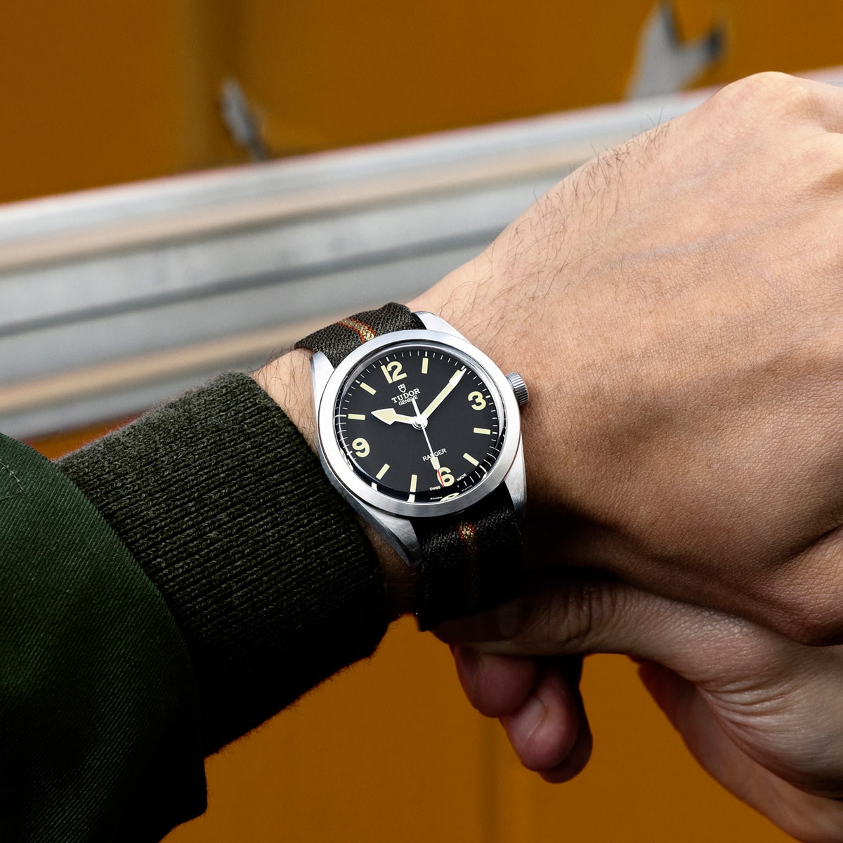 2025 © TUDOR Eine Person, die eine schwarz-silberne Tudor Ranger-Armbanduhr mit Stoffarmband und gelben Ziffern auf dem Zifferblatt trägt, hält ihr Handgelenk vor einem unscharfen orangefarbenen Hintergrund.