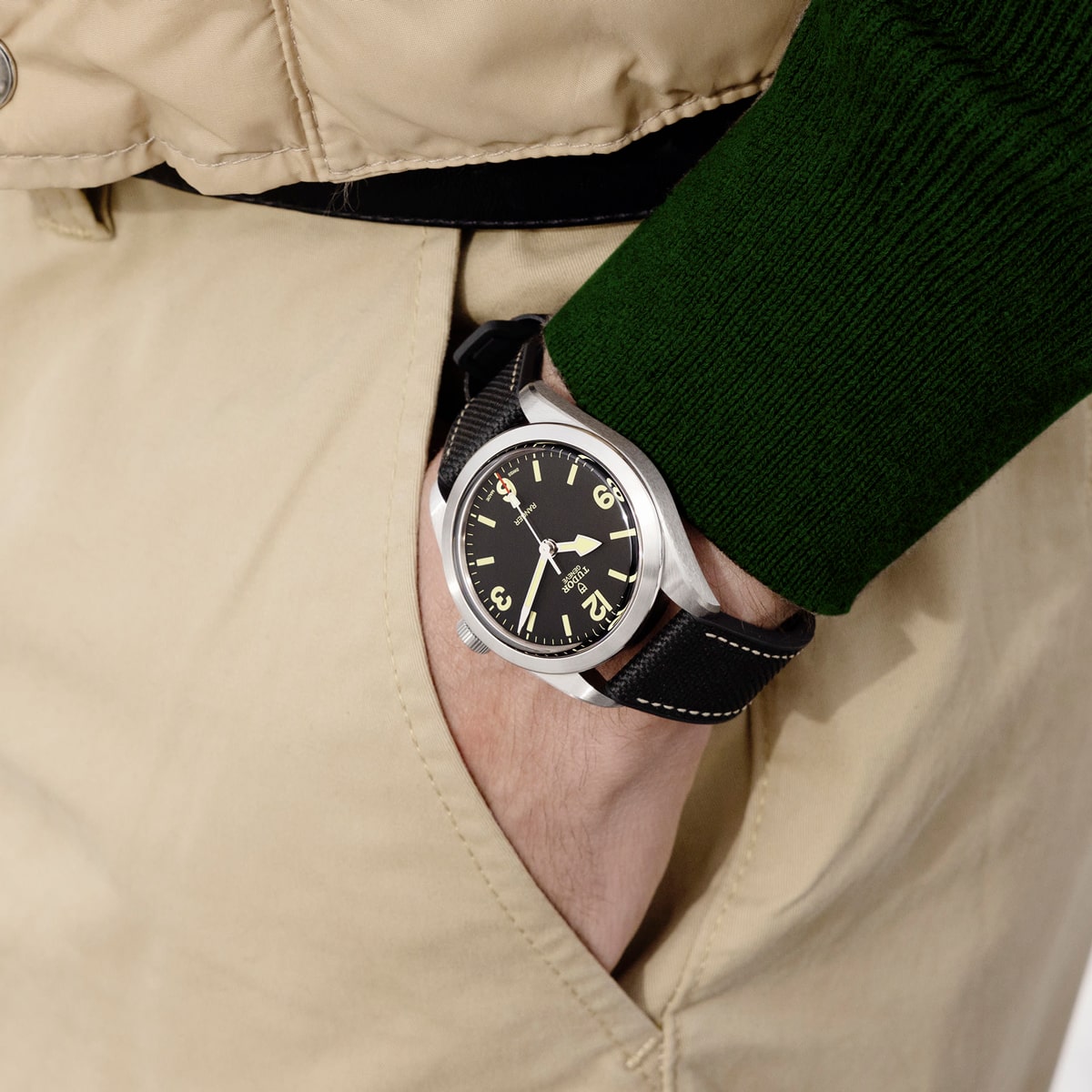 2025 © TUDOR Eine Person, die eine beige Hose und einen grünen Pullover trägt, hat die Hand in der Tasche und präsentiert eine schwarze Tudor Ranger Armbanduhr mit schwarzem Armband und großen gelben Zahlen auf dem Zifferblatt.