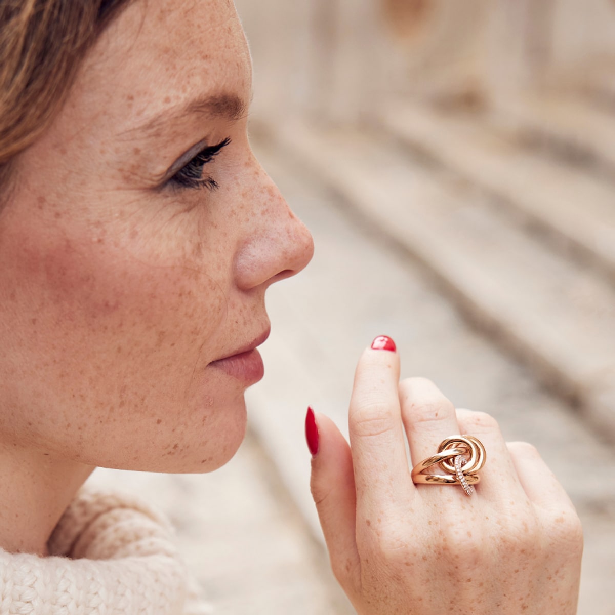 Seitenprofil einer Frau mit Sommersprossen und rotem Nagellack in Nahaufnahme, die einen Goldring von Capolavoro Ringe mit einem komplizierten Knotenmuster trägt. Sie blickt nachdenklich, mit verschwommenen Steinstufen im Hintergrund.