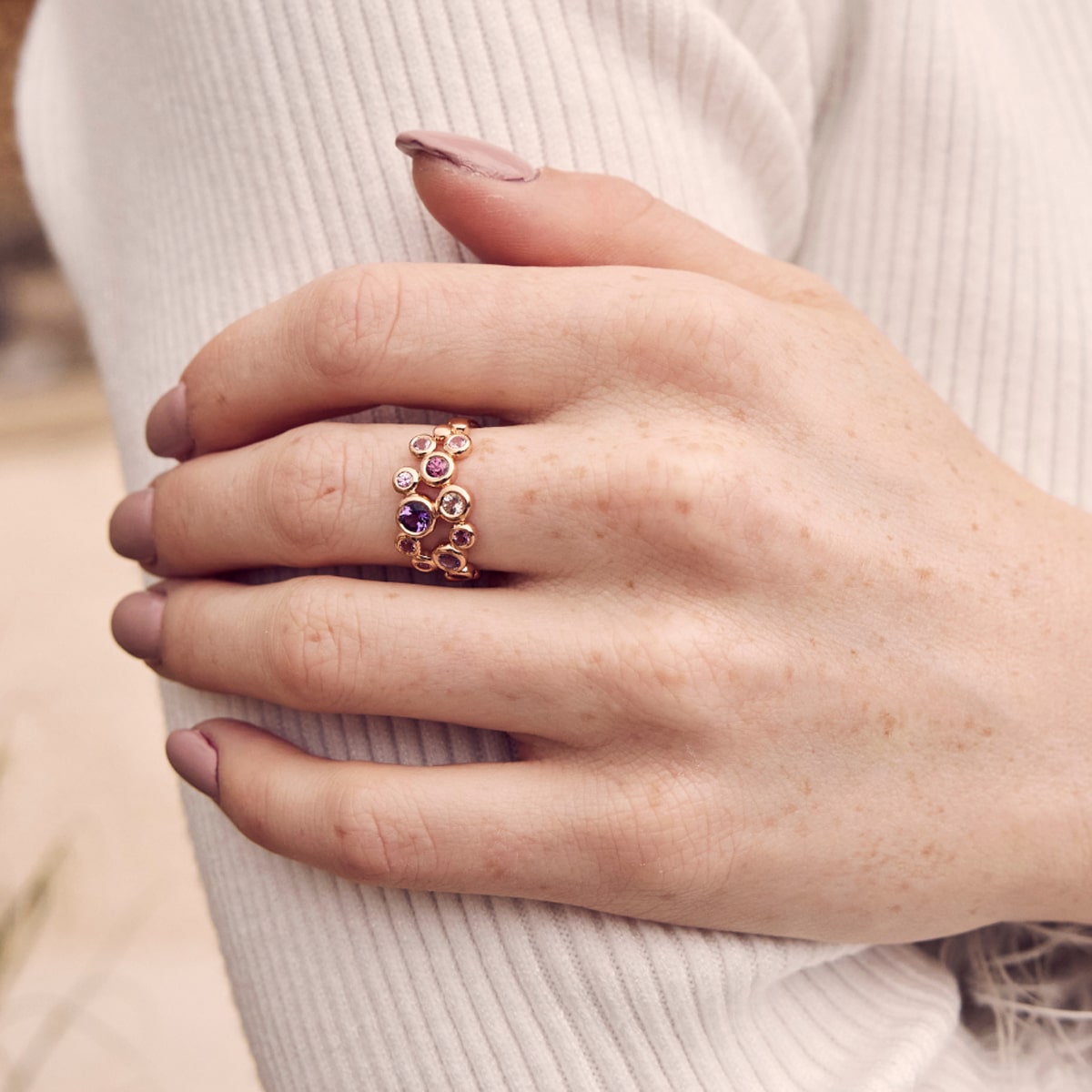 Nahaufnahme einer Hand mit heller Haut, die einen mit bunten Edelsteinen besetzten Goldring von Capolavoro Ringe trägt, der auf einem gerippten, cremefarbenen Ärmel ruht. Die Nägel sind manikürt und in einem Nude-Ton lackiert.