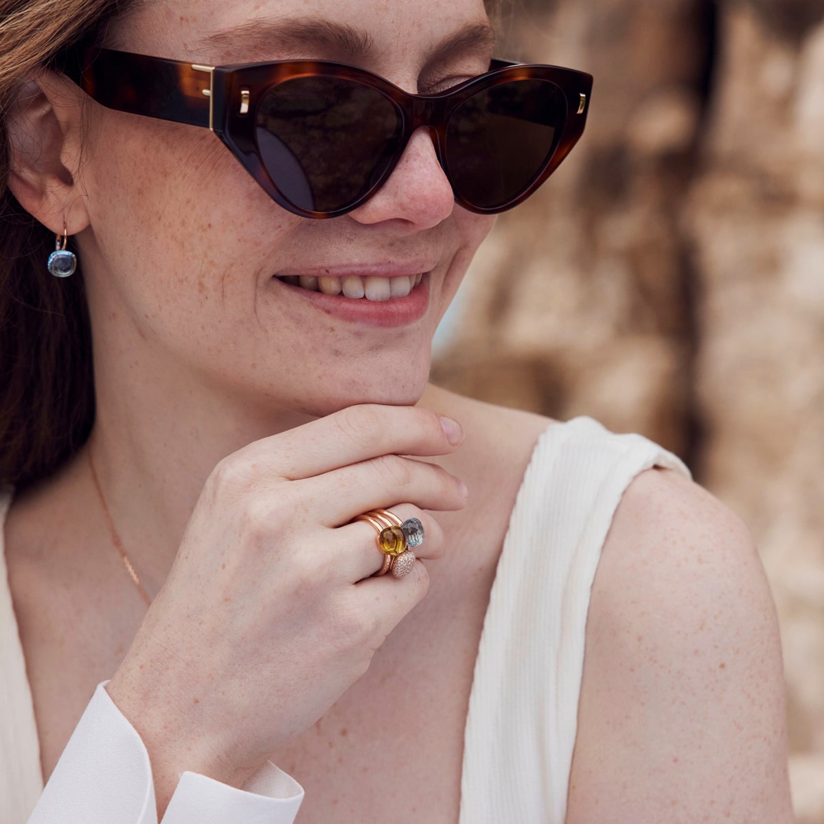 Eine Frau, die eine Schildpatt-Sonnenbrille und ein ärmelloses weißes Oberteil trägt, lächelt und zeigt mehrere Capolavoro Ringe und einen Ohrring. Sie hat helle Haut, Sommersprossen, und ihre Hand berührt sanft ihr Kinn.