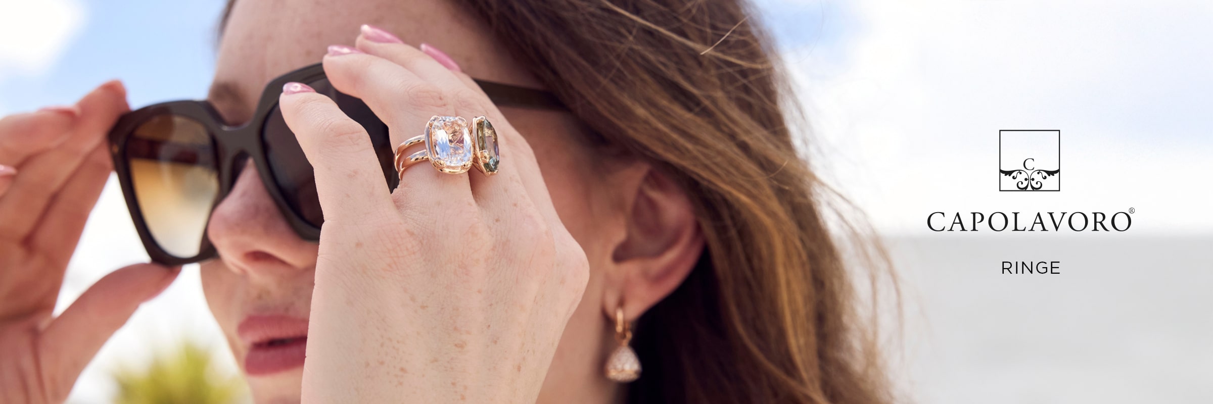 Eine Frau mit großer schwarzer Sonnenbrille, Edelsteinringen und goldenen Ohrringen steht im Freien im Sonnenlicht. Der Text neben ihr lautet "Capolavoro Ringe" und hebt die Eleganz des Schmucks von Capolavoro Ringe mit einem Logo darüber hervor.