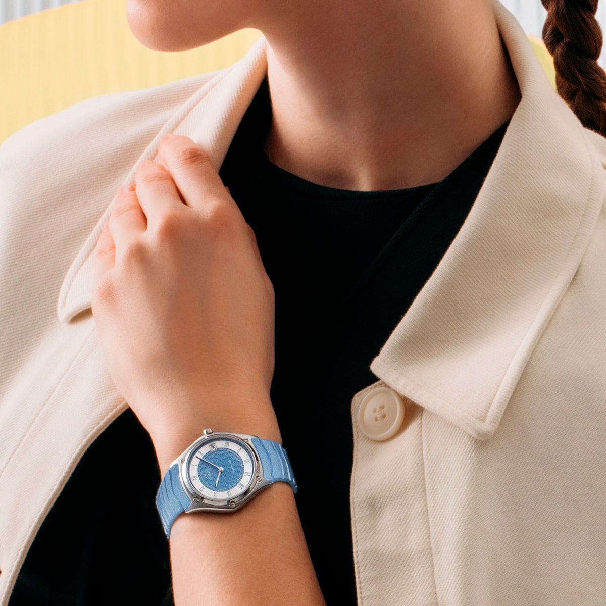 2025 © EBEL Eine Person, die eine helle Jacke und eine blaue EBEL Sport Classic Armbanduhr mit rundem Zifferblatt trägt, stellt ihren Kragen ein. Der Hintergrund weist weiche, verschwommene Farben auf und lenkt die Aufmerksamkeit auf die Uhr, die sie am Handgelenk trägt.