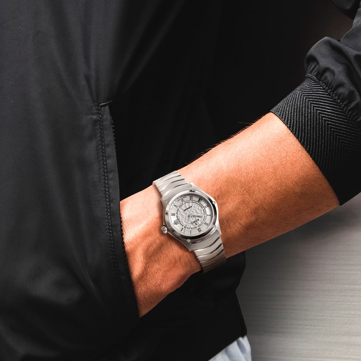 2025 © EBEL Eine Person, die eine schwarze Jacke trägt, hat die Hand in der Tasche und zeigt eine silberne EBEL Sport Classic Armbanduhr mit rundem Ziffernblatt und einem segmentierten Metallband.