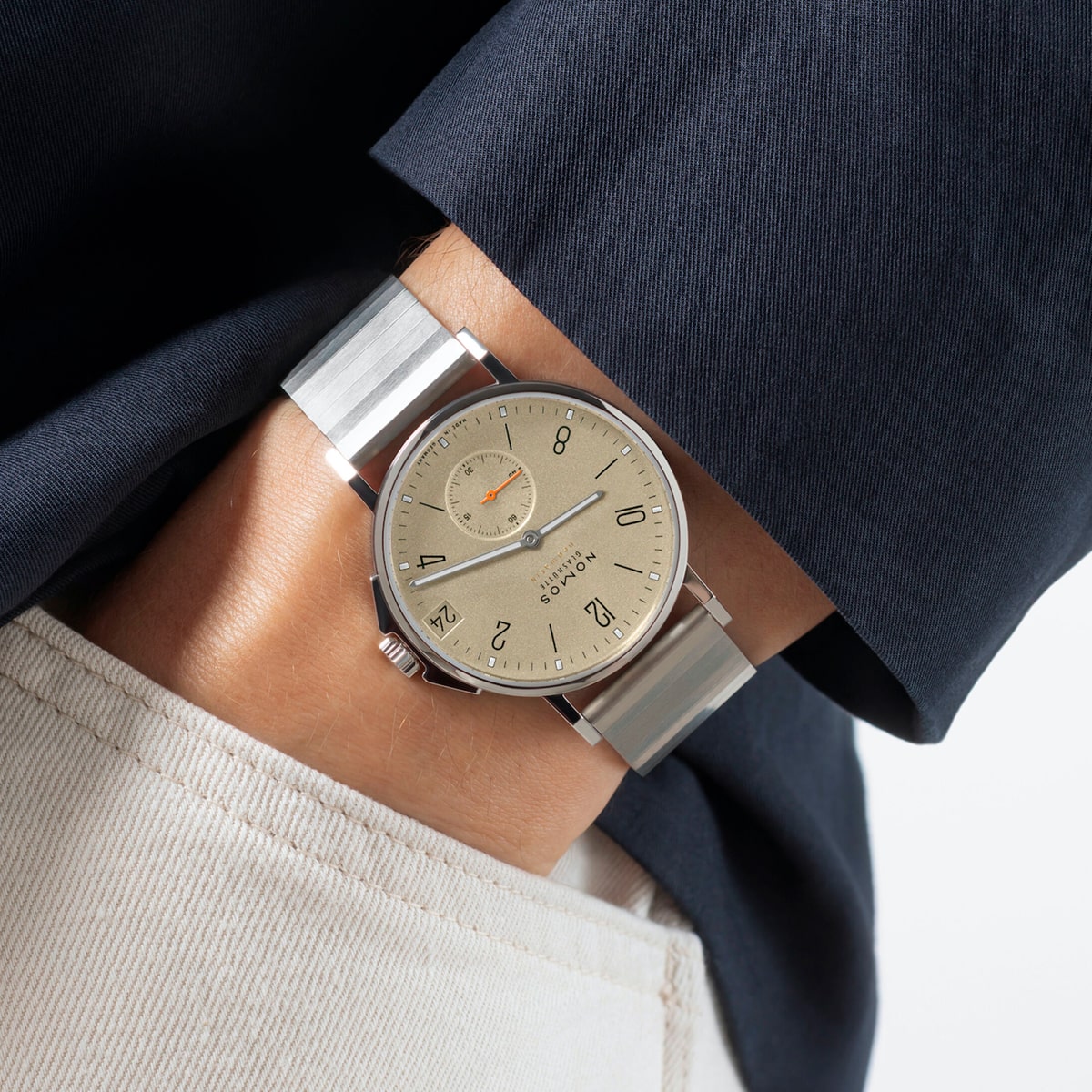 2025 © NOMOS GLASHÜTTE Nahaufnahme einer Person, die eine silberne NOMOS Glashütte Ahoi-Armbanduhr mit beigem Zifferblatt und schwarzen Ziffern trägt, bekleidet mit einem dunkelblauen Blazer und einer cremefarbenen Hose.
