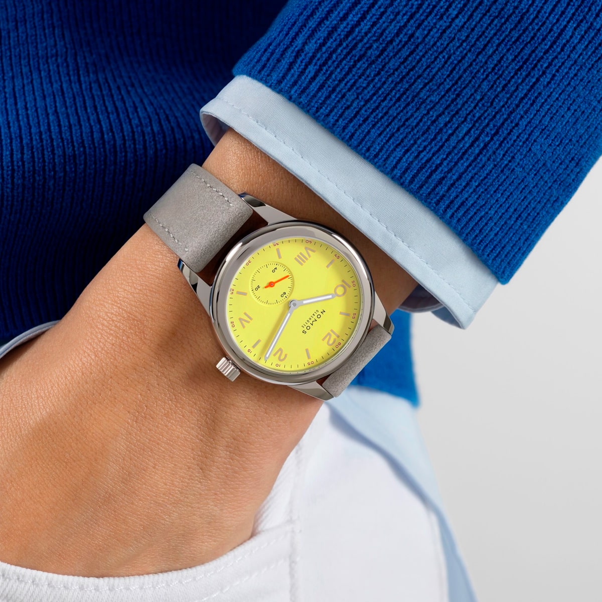 2025 © NOMOS GLASHÜTTE Eine Person, die einen blauen Pullover und ein hellblaues Hemd trägt, zeigt eine NOMOS Glashütte Club-Armbanduhr mit gelbem Zifferblatt, grauem Armband und roten Akzenten auf dem Ziffernblatt. Die Person ist außerdem mit einer weißen Hose bekleidet.