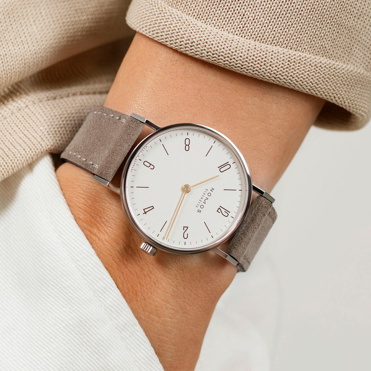Nahaufnahme eines Handgelenks mit einer minimalistischen silbernen NOMOS Glashütte Tangente mit weißem Zifferblatt, grauem Wildlederarmband und einfachen schwarzen Zahlen. Die Person ist mit einem beigen Oberteil und einem weißen Hemd bekleidet.