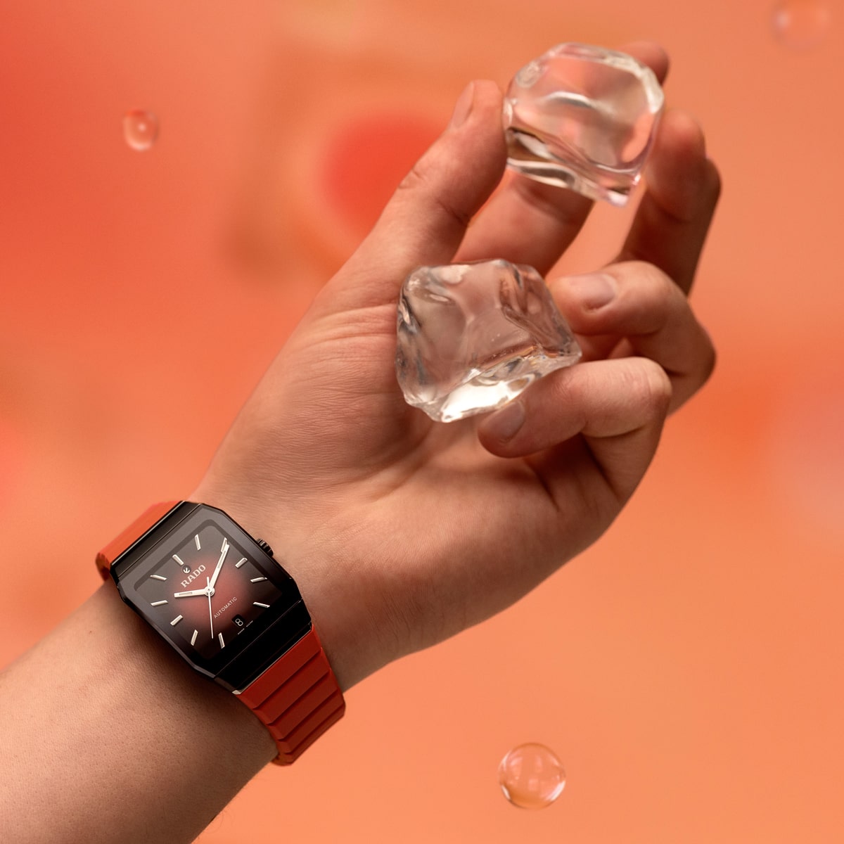 2025 © RADO Eine Hand, die eine rote Rado Anatom-Armbanduhr trägt, hält zwei klare Eiswürfel vor einem zart orangefarbenen Hintergrund, auf dem einige Wassertropfen zu sehen sind.