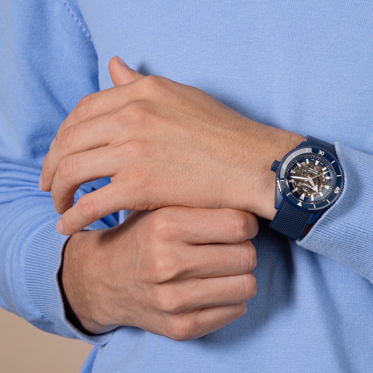 2025 © RADO Eine Person, die einen hellblauen Pullover trägt, stellt eine Rado Captain Cook mit einem sichtbaren mechanischen Uhrwerk an ihrem linken Handgelenk ein. Die Uhr hat ein strukturiertes blaues Armband und ein rundes Zifferblatt.