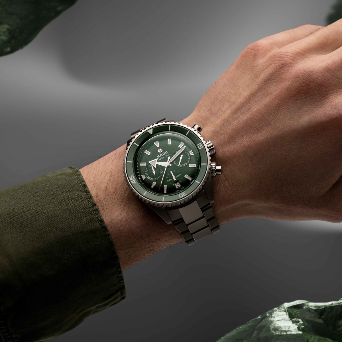 2025 © RADO Nahaufnahme einer Person, die eine silberne Rado Captain Cook Metallarmbanduhr mit grünem Zifferblatt und mehreren Hilfszifferblättern trägt. Der olivgrüne Ärmel der Person ergänzt den weichen, verschwommenen Farbverlauf mit grünen Akzenten im Hintergrund.