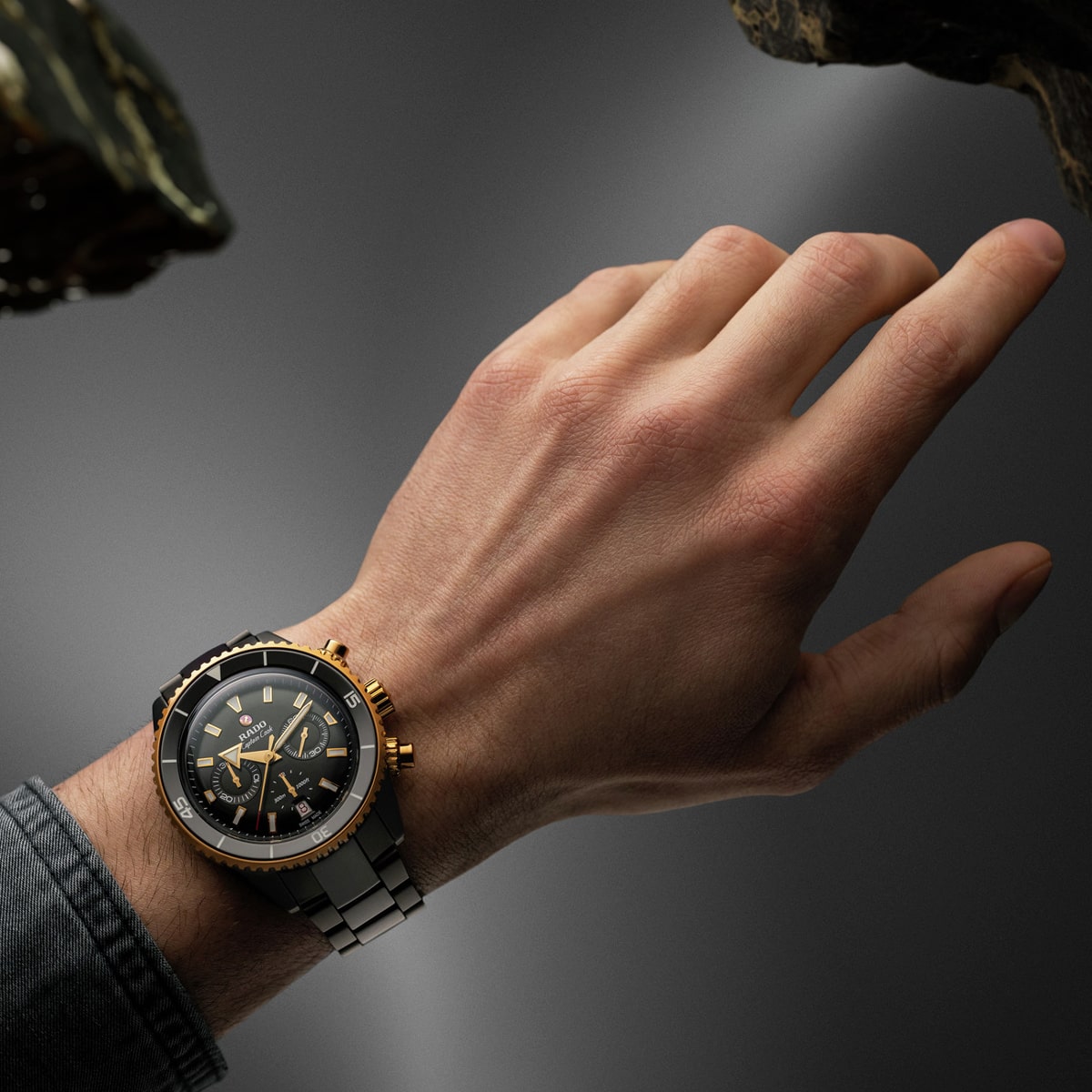 2025 © RADO Eine Nahaufnahme der linken Hand einer Person, die eine luxuriöse silberne und goldene Rado Captain Cook Armbanduhr mit schwarzem Zifferblatt trägt, vor einem dunklen, schwach beleuchteten Hintergrund. Die Person ist mit einem grauen Ärmel bekleidet.