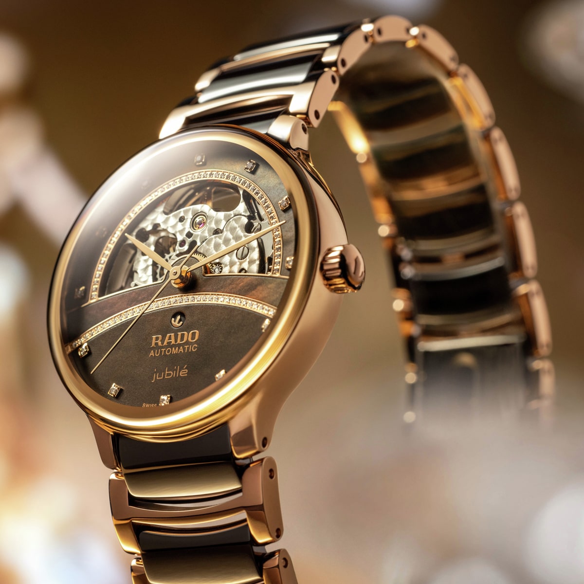 2025 © RADO Eine Nahaufnahme einer luxuriösen Rado Centrix Jubile Automatik-Armbanduhr mit goldenem und schwarzem Metallband, mit teilweise skelettiertem Zifferblatt und Diamantenakzenten, vor einem sanft verschwommenen Hintergrund.