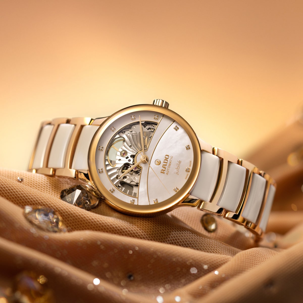 2025 © RADO Eine goldene und weiße Rado Centrix Luxus-Armbanduhr mit einem teilweise geöffneten Zifferblatt, das den Blick auf die innere Mechanik freigibt, wird elegant auf einem funkelnden, goldfarbenen Stoff vor einem warmen, sanft beleuchteten Hintergrund präsentiert.