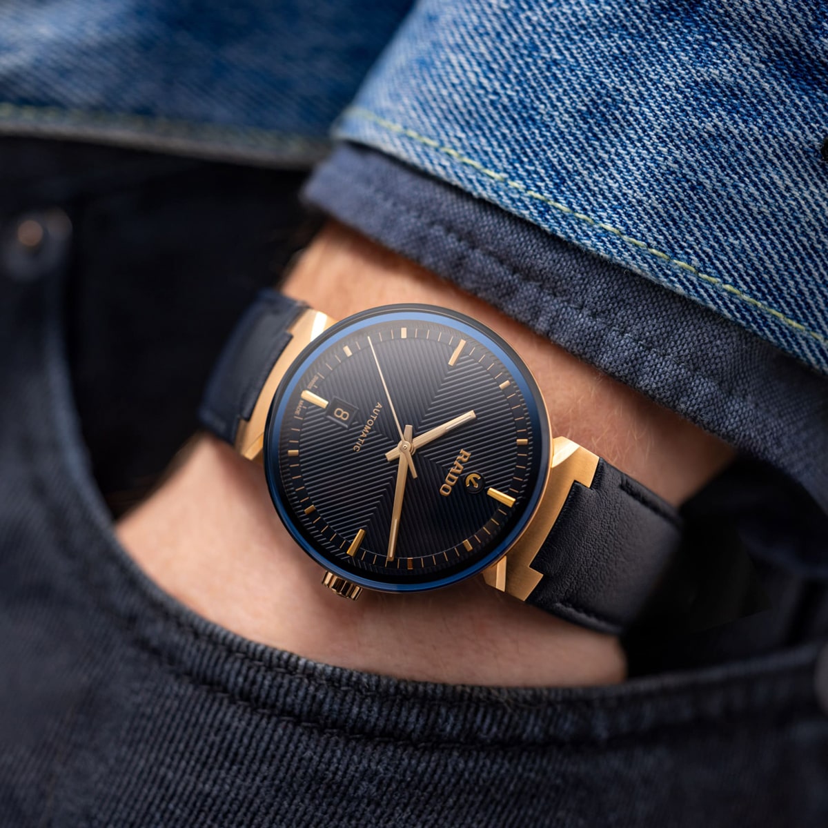 2025 © RADO Eine Nahaufnahme des Handgelenks einer Person, die eine elegante schwarz-goldene Uhr von Rado Florence trägt, die teilweise in der Tasche einer dunkelblauen Jeans und unter einer blauen Jeansjacke steckt.