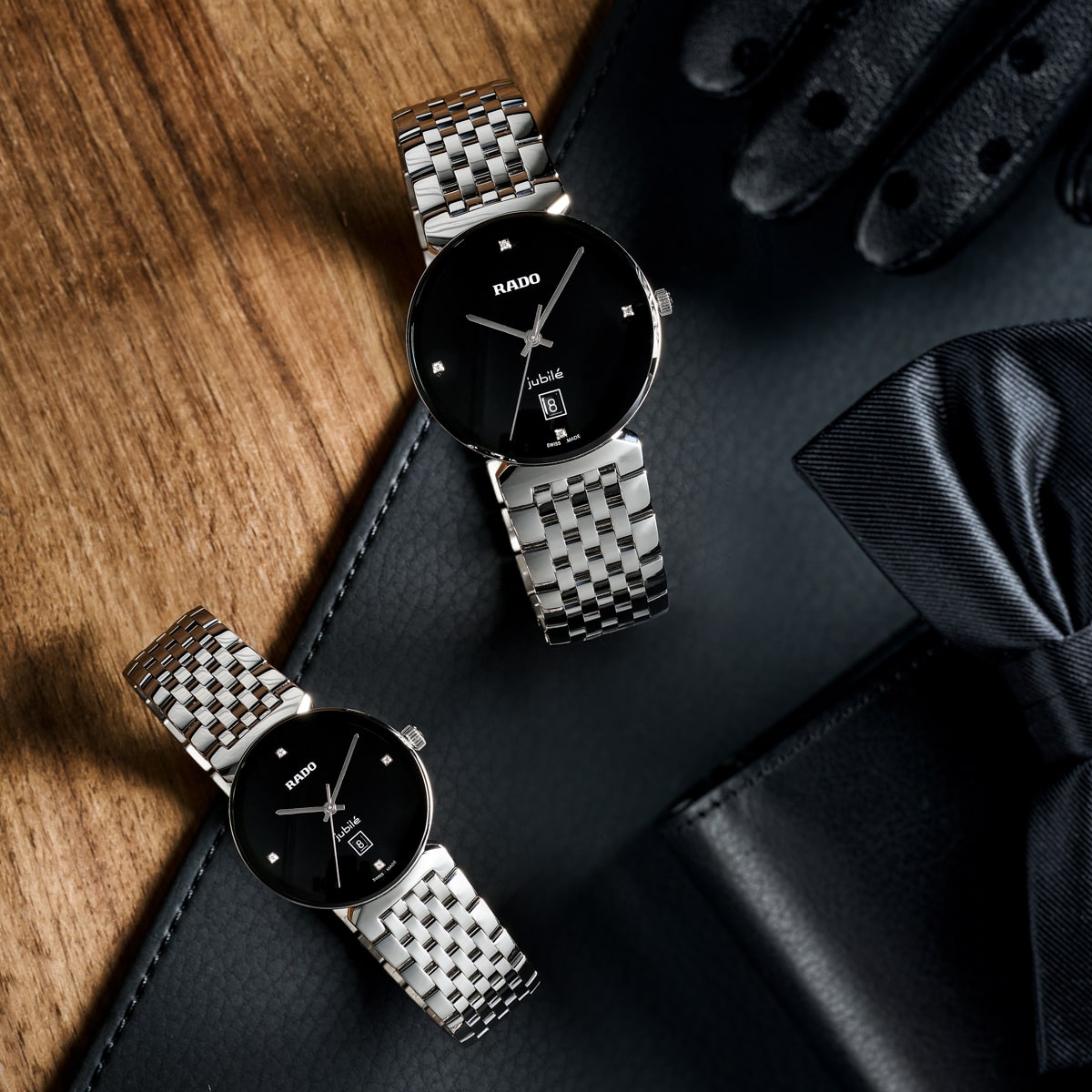 2025 © RADO Zwei elegante silberne Rado Florence Uhren mit schwarzen Ziffernblättern liegen auf einer Holzfläche neben schwarzen Lederhandschuhen, einer schwarzen Fliege und einer schwarzen Brieftasche. Die Szene suggeriert einen anspruchsvollen, formellen Stil.