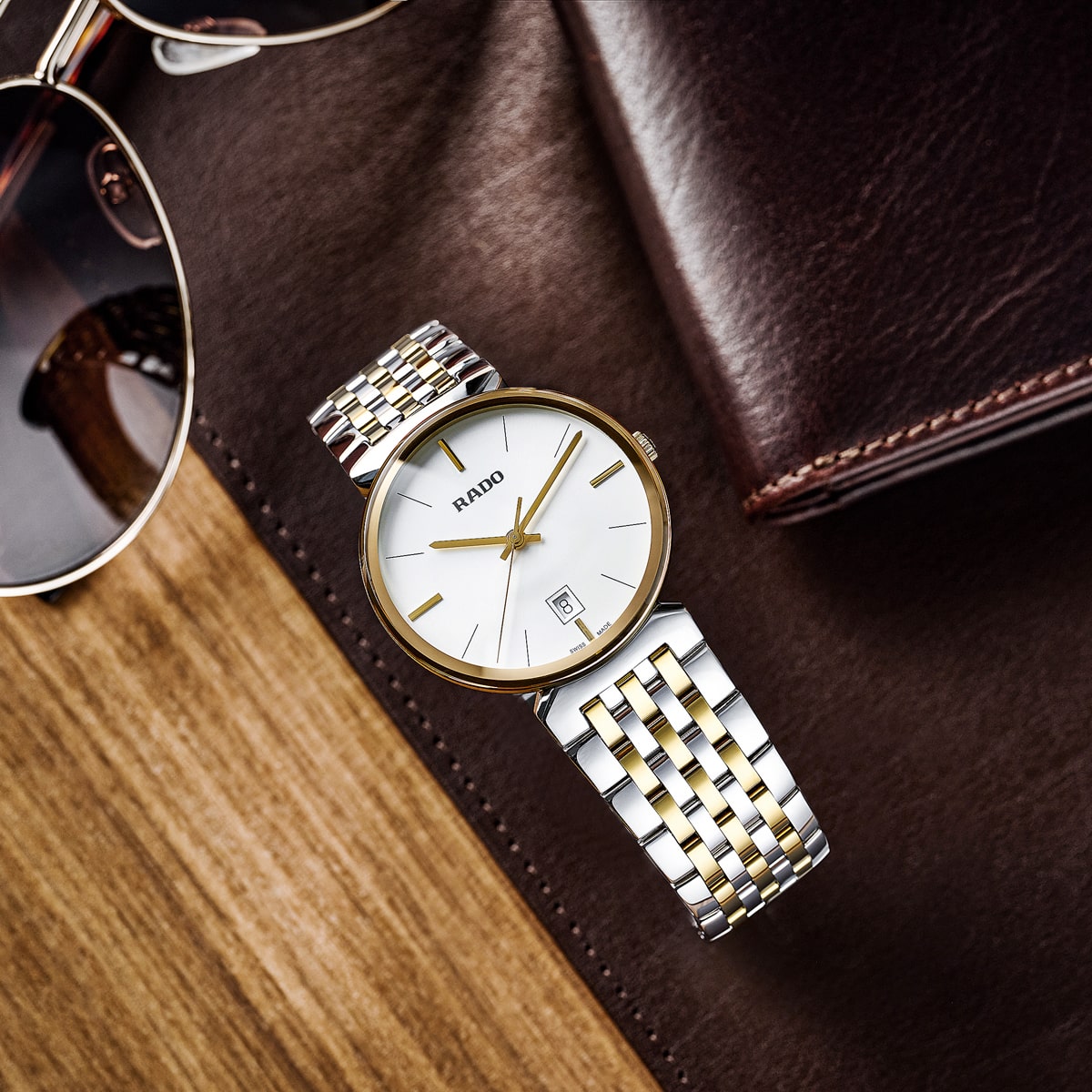 2025 © RADO Eine silber- und goldfarbene Rado Florence-Armbanduhr mit weißem Zifferblatt liegt auf einer braunen Lederfläche neben einer Piloten-Sonnenbrille und einem Leder-Notizbuch, die alle elegant auf einem Holztisch angeordnet sind.
