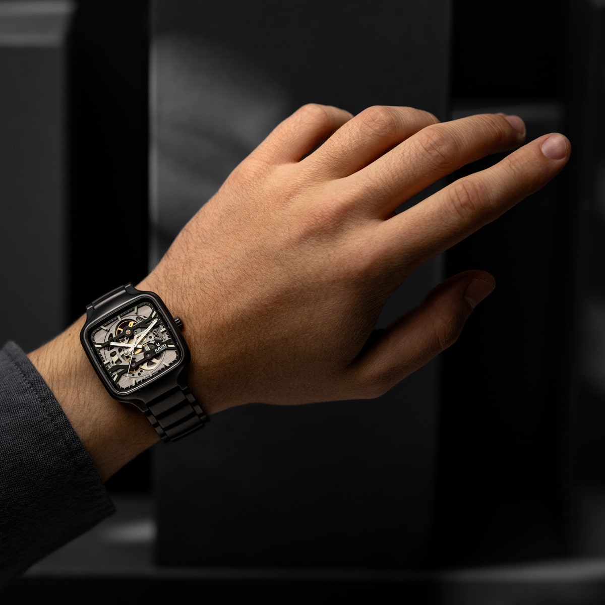 2025 © RADO Nahaufnahme einer linken Hand, die eine skelettierte Rado True Square Uhr aus schwarzem Metall trägt, die ein kompliziertes Innenleben zeigt, vor einem dunklen, unscharfen Hintergrund. Die Person ist mit einem dunklen langärmeligen Hemd bekleidet.