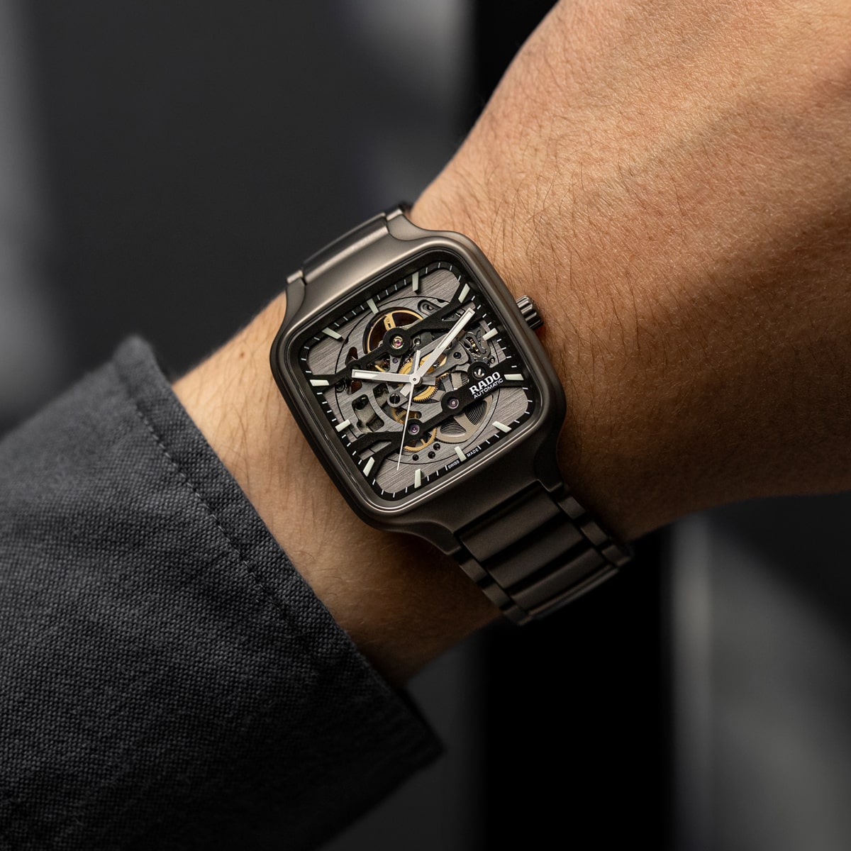 2025 © RADO Eine Person, die eine metallene Rado True Square Skelettuhr mit sichtbaren Zahnrädern und einem schwarzen Armband trägt, bekleidet mit einem dunklen langärmeligen Hemd.