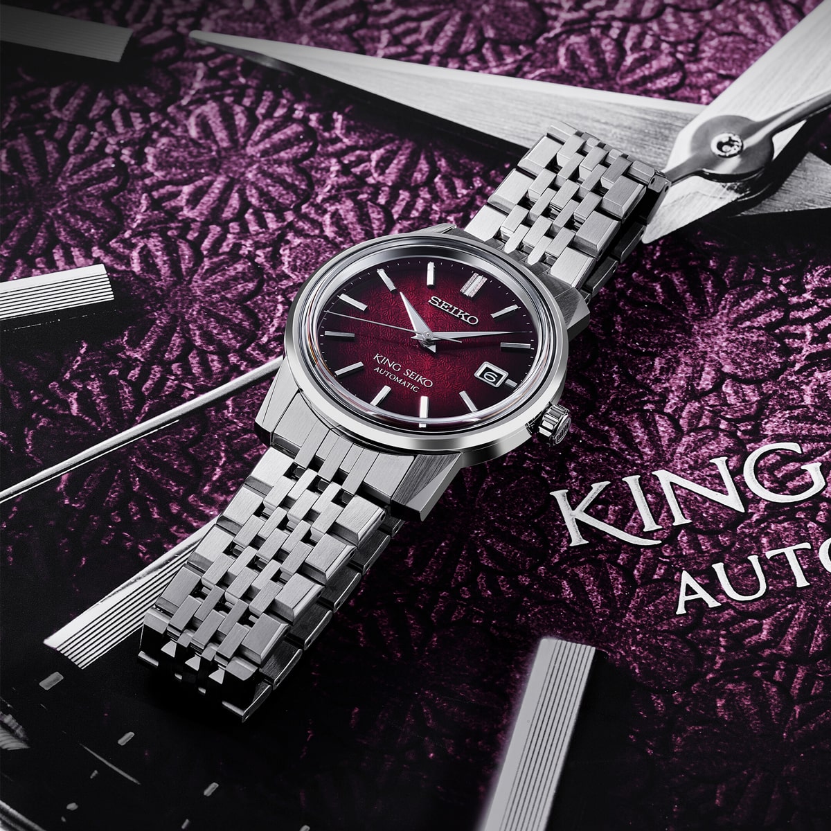 Eine King Seiko-Armbanduhr mit silbernem Metallarmband und tiefrotem Zifferblatt ruht auf einem violett gemusterten Hintergrund, der mit großen Zeigern und Text verziert ist.