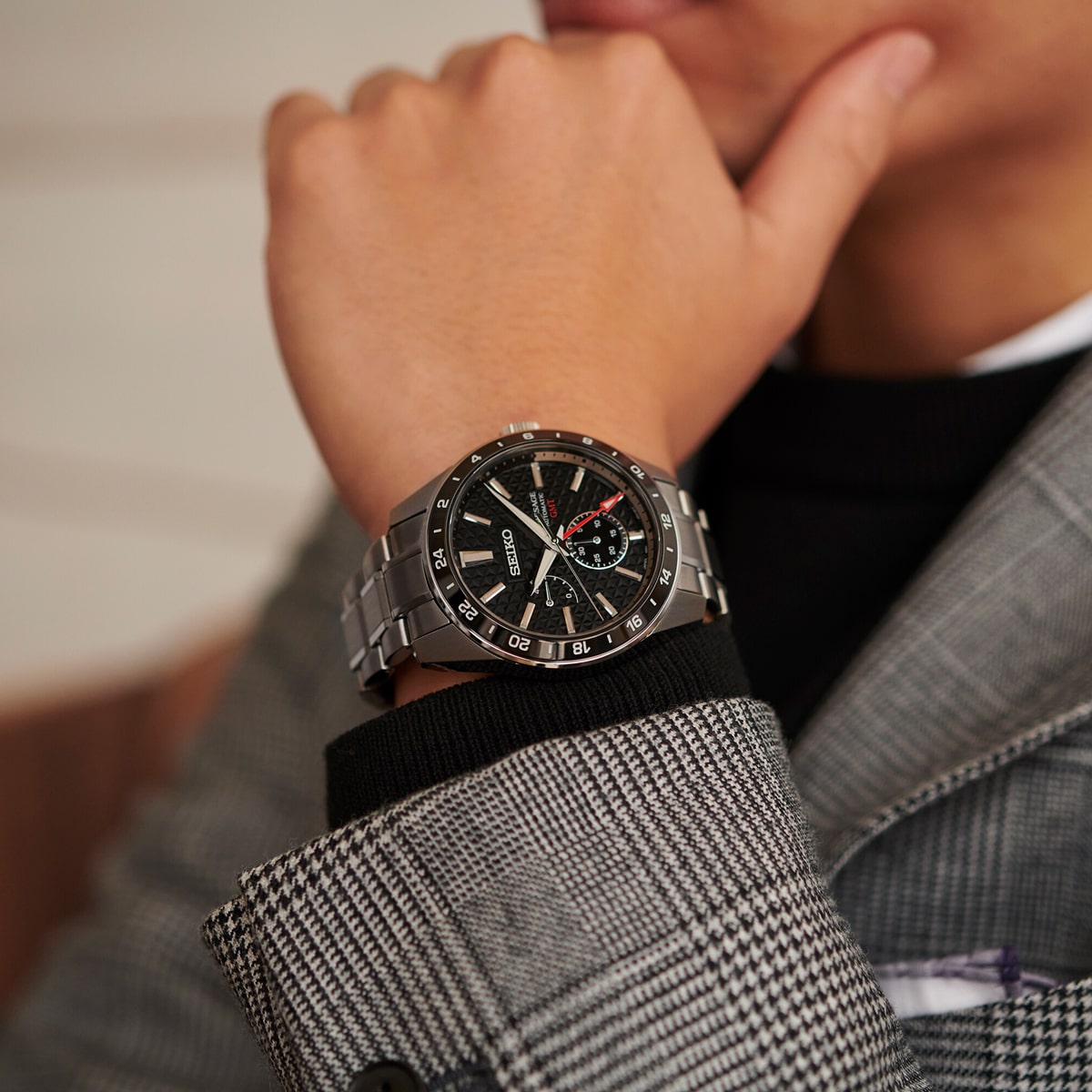 Eine Person in einem grau karierten Anzug und einem schwarzen Rollkragenpullover stützt ihr Kinn auf die Hand und präsentiert eine Seiko Presage Armbanduhr mit silbernem Gehäuse, schwarzem Zifferblatt und auffälligen roten Akzenten.