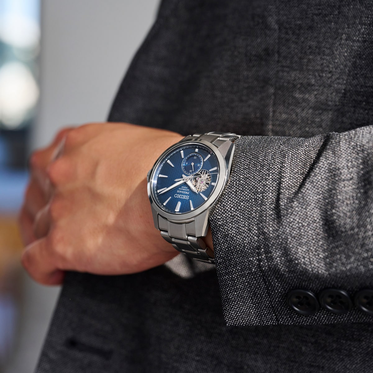 Nahaufnahme einer Person, die eine graue Anzugsjacke und eine Seiko Presage trägt, mit einer silbernen Armbanduhr mit blauem Zifferblatt und sichtbarem Innenleben, die prominent am linken Handgelenk getragen wird.