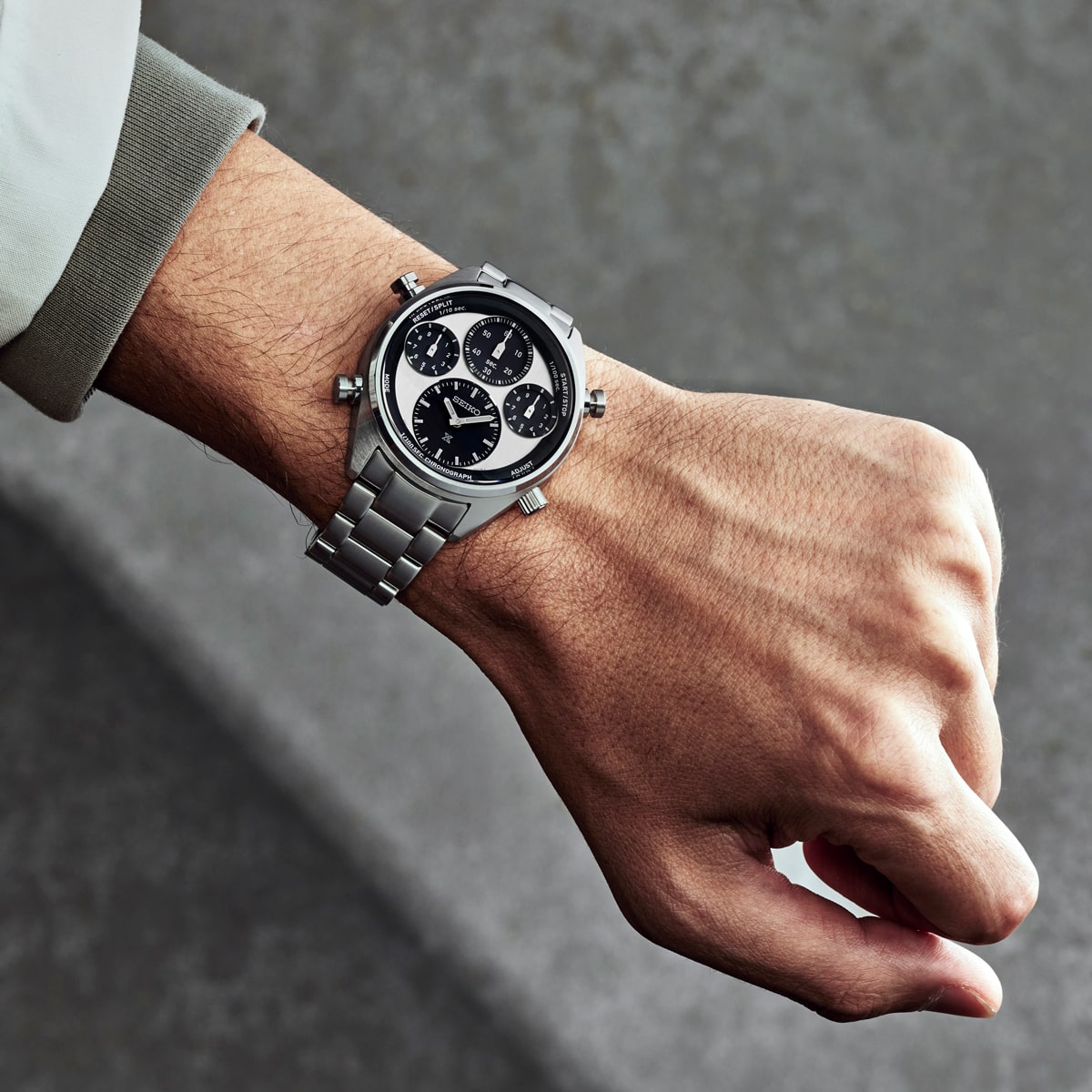 Nahaufnahme des linken Handgelenks einer Person, die eine Seiko Prospex Edelstahl-Chronographenuhr mit schwarzem Zifferblatt und drei Unterzifferblättern trägt, vor einem unscharfen grauen Hintergrund.