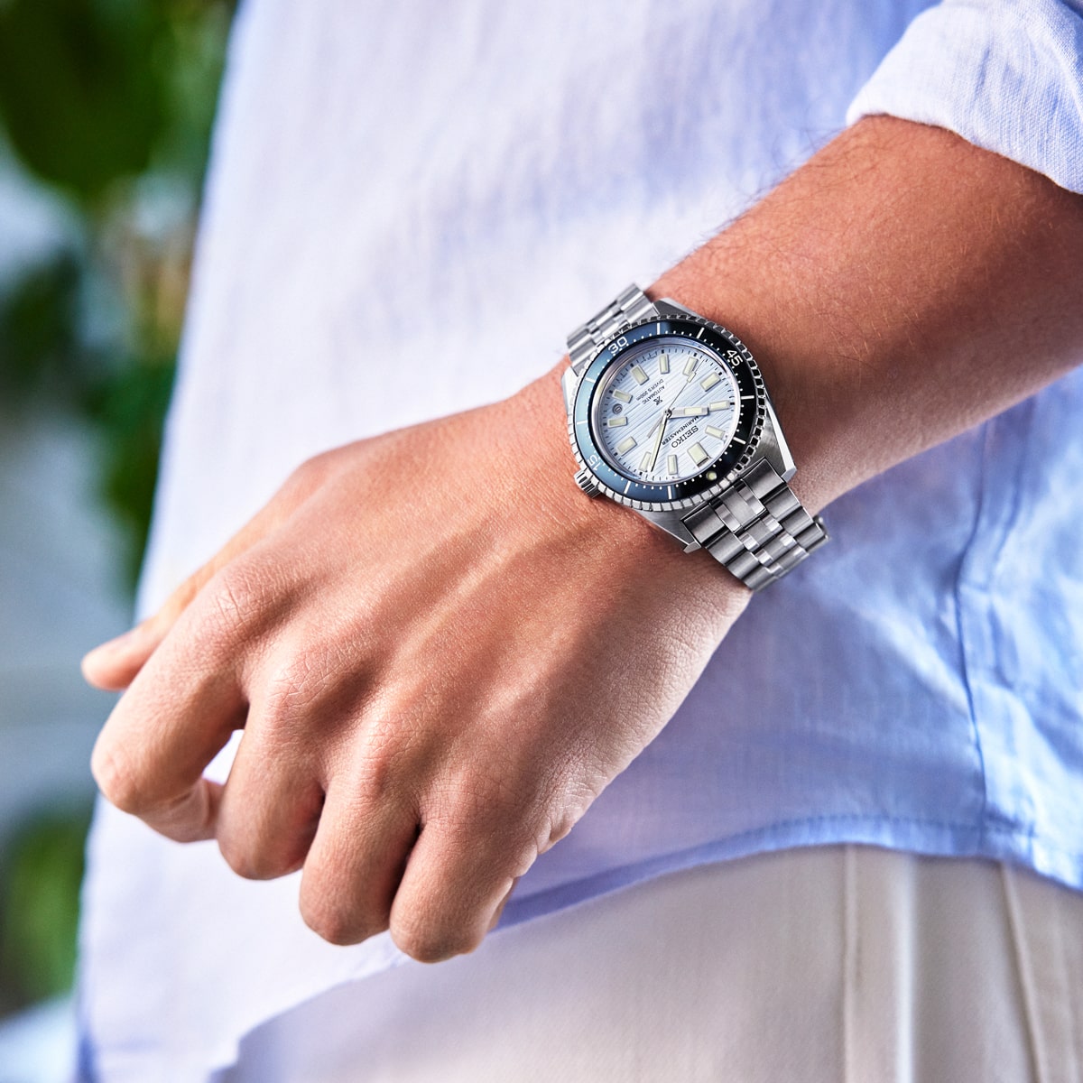 Eine Person trägt eine silberne Seiko Prospex-Armbanduhr mit Metallband und weißem Zifferblatt. Die Person ist mit einem hellblauen langärmeligen Hemd und einer beigen Hose bekleidet, im Hintergrund ist unscharfes Grün zu sehen.