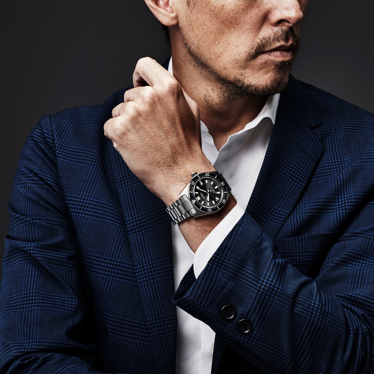 Ein Mann in einem blau karierten Anzug und weißem Hemd zeigt eine silberne Seiko Prospex-Armbanduhr mit schwarzem Zifferblatt und verschränkt vor einem dunklen Hintergrund den Arm vor der Brust.