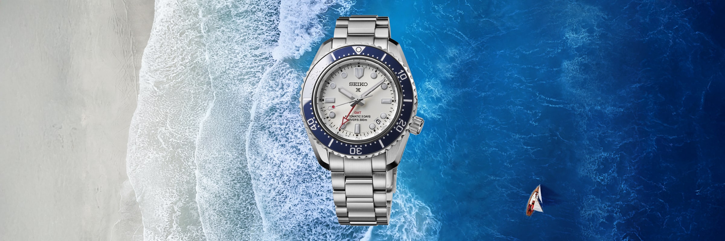 Eine silberne Seiko Prospex Taucheruhr mit blauer Lünette ist über einer Luftaufnahme eines Sandstrandes, weißer Wellen und eines kleinen Segelbootes, das im tiefblauen Meer treibt, zentriert.