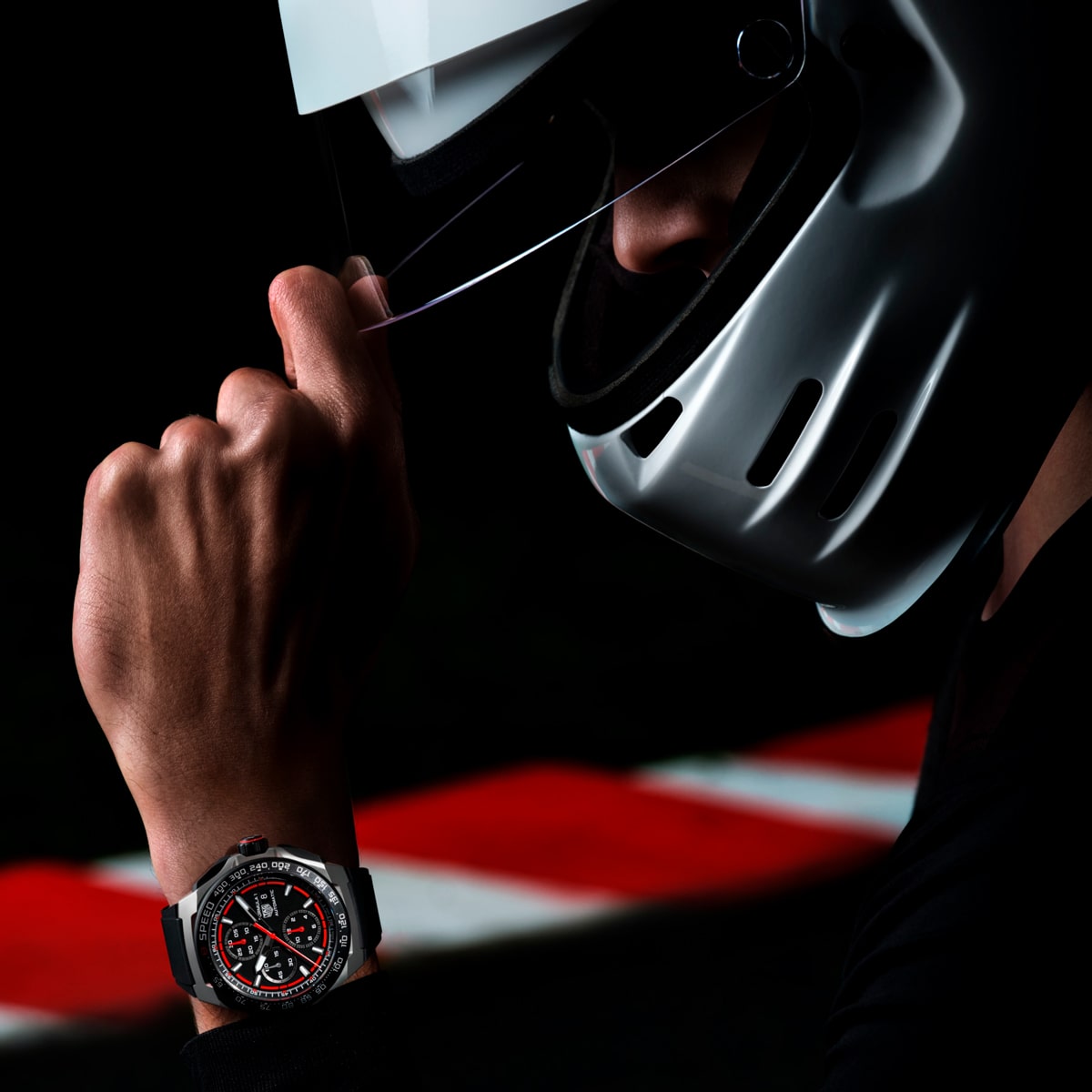 Eine Person, die einen weißen Rennhelm trägt, klappt das Visier hoch und enthüllt einen Teil ihres Gesichts. An ihrem Handgelenk glänzt eine elegante schwarz-silberne TAG Heuer Formel-1-Sportuhr, im Hintergrund verschwimmt eine Rennstrecke.