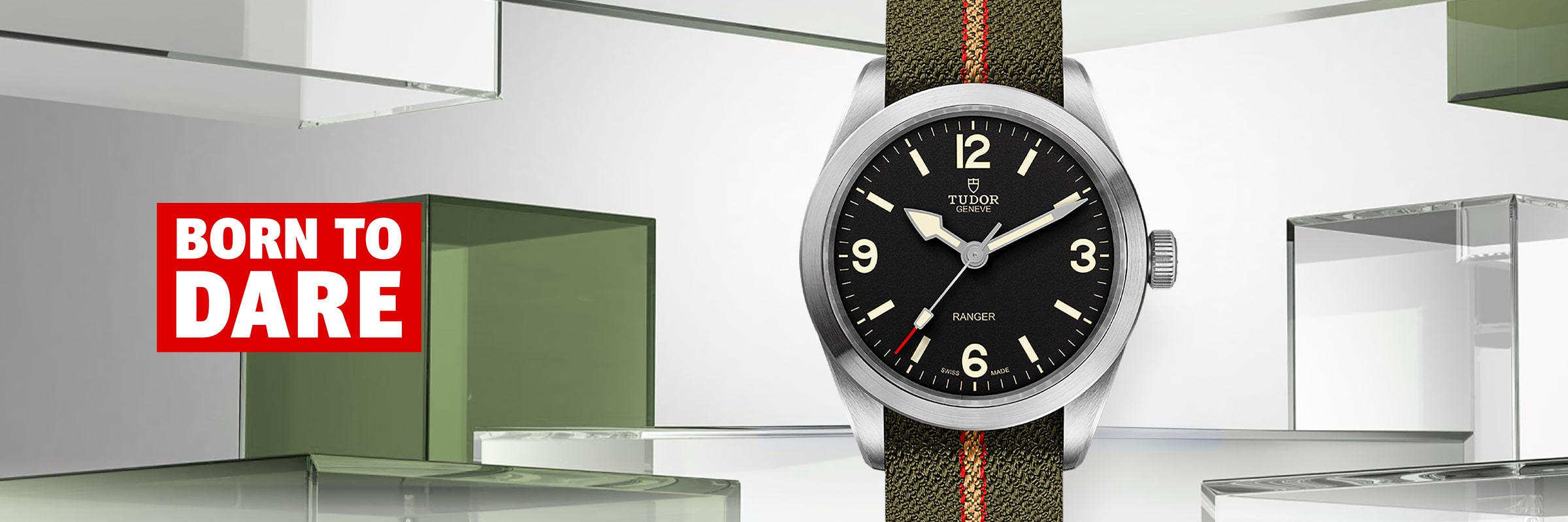 2025 © TUDOR Eine Tudor Ranger Armbanduhr mit grünem Armband und schwarzem Zifferblatt steht neben dem plakativen rot-weißen Slogan "BORN TO DARE", der von geometrischen Glasformen im Hintergrund akzentuiert wird.