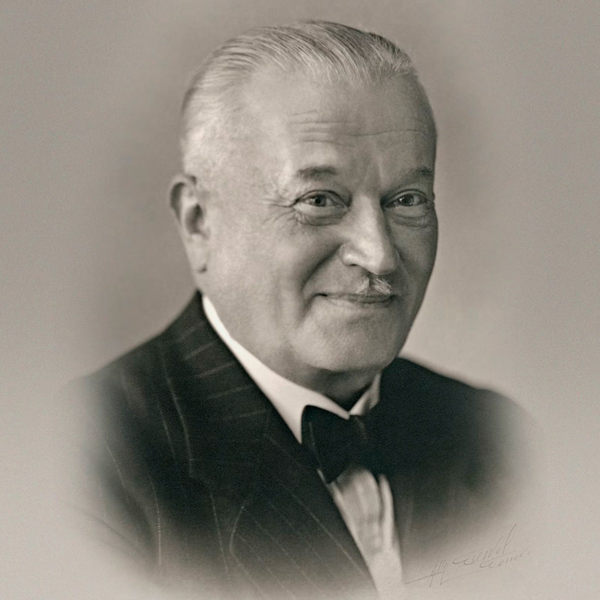 Ein älterer Mann mit hellem, zurückgekämmtem Haar, der einen dunklen Nadelstreifenanzug im Tudor-Stil, ein weißes Hemd und eine Fliege trägt, lächelt sanft in diesem Schwarz-Weiß-Porträt mit weichem, unscharfem Rand.