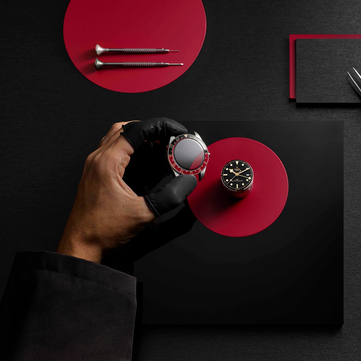 Eine behandschuhte Hand hält eine Tudor-Uhrenlünette neben einer schwarzen Armbanduhr auf einem roten und schwarzen geometrischen Hintergrund, flankiert von zwei Schraubenziehern und einer Pinzette.
