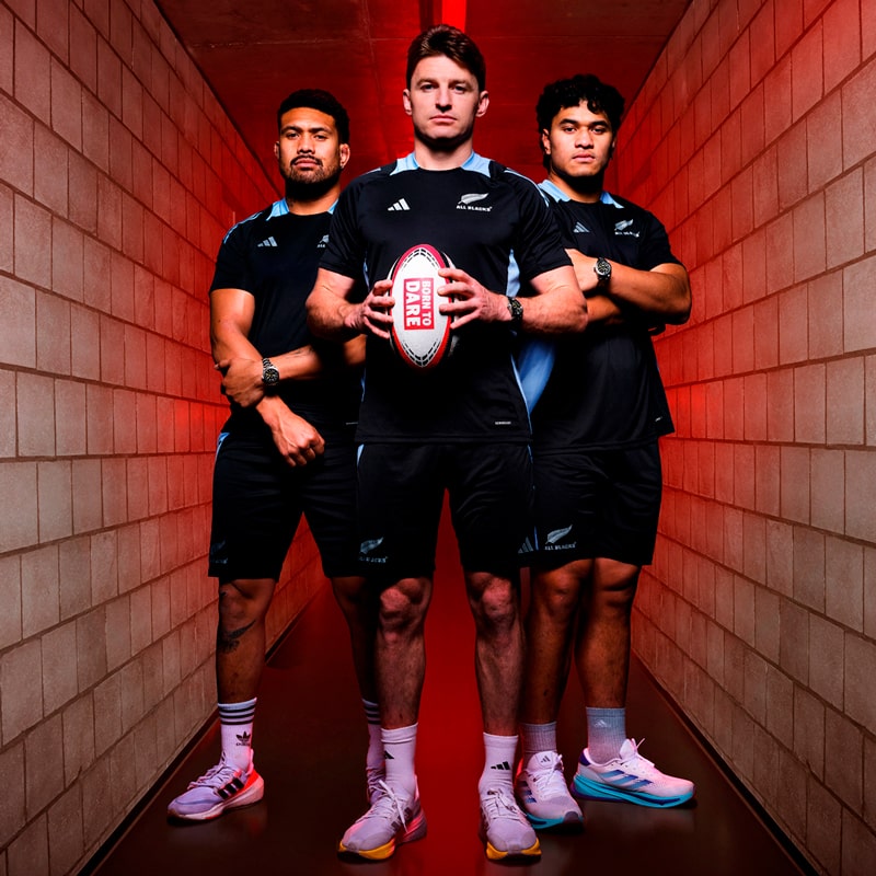 Drei männliche Rugbyspieler in schwarzen Uniformen stehen selbstbewusst in einem Tunnel mit roter Beleuchtung, der mittlere Spieler hält einen Rugbyball. Mit ernster Miene und Tudor-ähnlicher Entschlossenheit bereiten sie sich auf den Kampf vor.