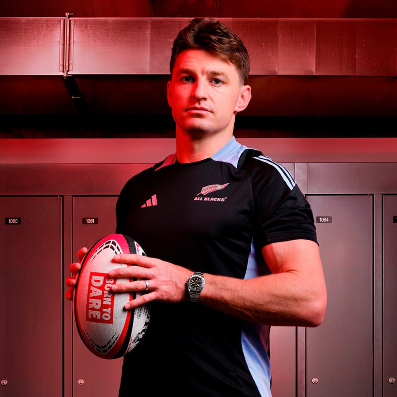 Ein männlicher Rugbyspieler im Trikot der All Blacks steht vor Umkleidekabinen und hält einen Rugbyball der Marke Tudor mit der Aufschrift "DARE TO DARE" in der Hand. Die rote Beleuchtung sorgt für einen dramatischen Effekt.