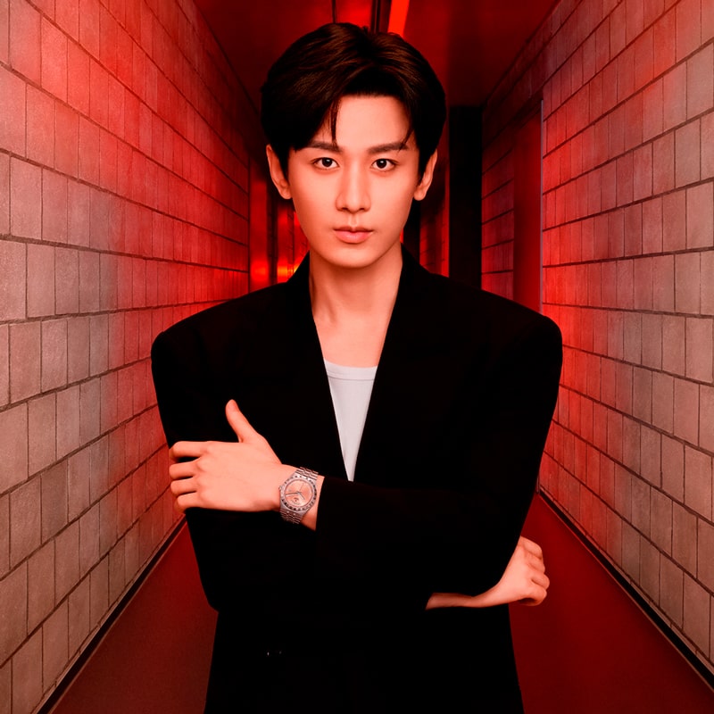 Ein junger Mann mit kurzen dunklen Haaren steht in einem schmalen Flur mit roter Beleuchtung und trägt einen schwarzen Blazer über einem weißen Hemd. Er verschränkt die Arme und blickt direkt in die Kamera, wobei er eine silberne Tudor-Armbanduhr zur Schau stellt.